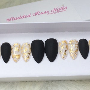 Puede incluir: Un conjunto de diez uñas postizas negras de stiletto con detalles de pan de oro. Las uñas se muestran en una caja blanca con el texto "Studded Rose Nails" y "Nails by Marquita".