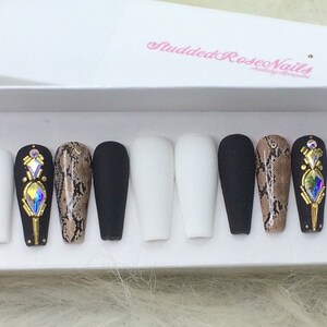 May include: Set of ten long, artificial nails with a variety of designs.  The nails are white, black, and brown with a snake skin pattern.  Some nails have gold accents and rhinestones.  The nails are displayed in a white box with the text "Studded Rose Nails" in pink.