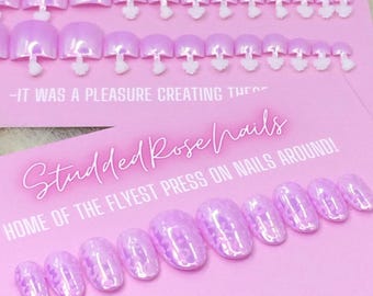 Puede incluir: Dos filas de uñas postizas de color lila claro con detalles blancos. Las uñas están dispuestas en una tarjeta rosa con el texto "Studded Rose nails" y "Home of the flyest press on nails around!"