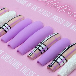 May include: Set of ten long, lavender-coloured press-on nails with a plaid design in black, white, and pink. The nails have a matte finish and are arranged on a pink background.