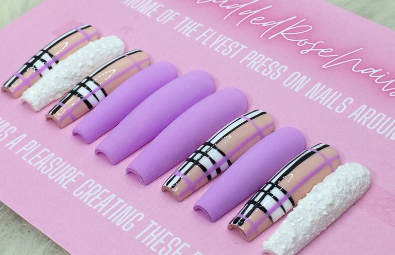 May include: Set of ten long, lavender-coloured press-on nails with a plaid design in black, white, and pink. The nails have a matte finish and are arranged on a pink background.