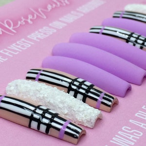May include: A set of long, light purple press-on nails with a black and white plaid design. Some of the nails have a white glitter accent. The nails are displayed on a pink surface with the text "The Flyest Press On Nails Around" and "It Was A Vibe".