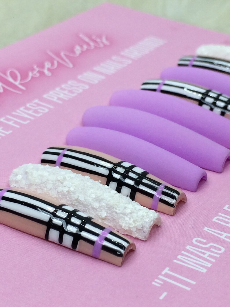 May include: A set of long, light purple press-on nails with a black and white plaid design. Some of the nails have a white glitter accent. The nails are displayed on a pink surface with the text "The Flyest Press On Nails Around" and "It Was A Vibe".