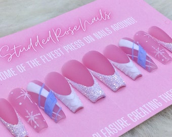 Puede incluir: Un conjunto de uñas postizas rosas con diseños de copos de nieve blancos y azules. Las uñas son largas y tienen un acabado mate. El texto "Studded Rose Nails" y "Home of the Flyest Press On Nails Around!" es visible en el fondo rosa.