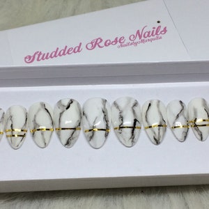 May include: A set of ten oval-shaped false nails with a white marble design and a gold accent stripe. The nails are displayed in a white box with the text "Studded Rose Nails" and "NailsbyMarquita".