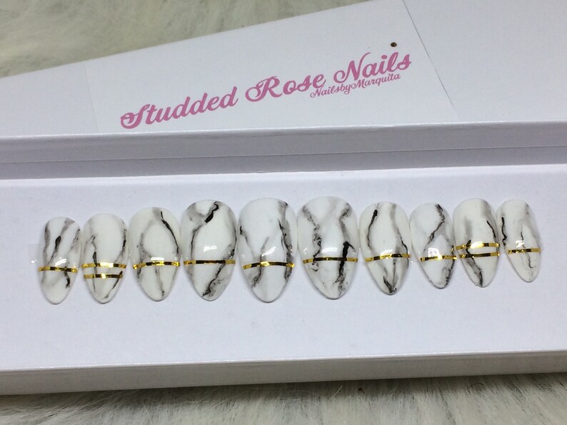 May include: A set of ten oval-shaped false nails with a white marble design and a gold accent stripe. The nails are displayed in a white box with the text "Studded Rose Nails" and "NailsbyMarquita".