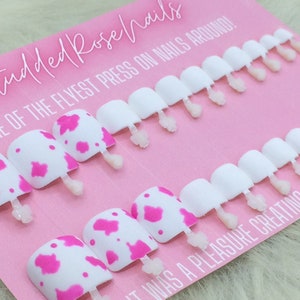 May include: A set of 10 press-on nails with a pink and white cow print design. The nails are displayed on a pink card with the text "Studded Rose Nails" and "Home of the Flyest Press On Nails Around!"