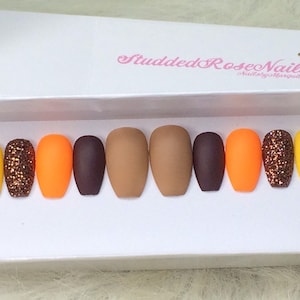 May include: A set of ten false nails in various colors, including yellow, orange, brown, and glitter. The nails are arranged in a row on a white surface. The text "Studded Rose Nails" is visible in the background.