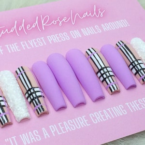 May include: A set of ten lavender press-on nails with a pink and black plaid design. The nails are long and have a matte finish. The text "Studded Rose Nails" and "Some of the flyest press on nails around!" is visible on the pink background.