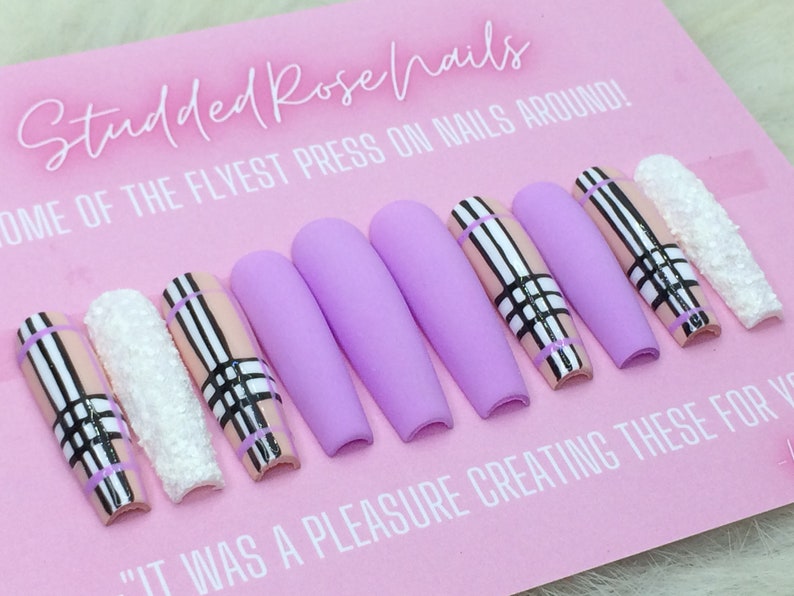 May include: A set of ten lavender press-on nails with a pink and black plaid design. The nails are long and have a matte finish. The text "Studded Rose Nails" and "Some of the flyest press on nails around!" is visible on the pink background.