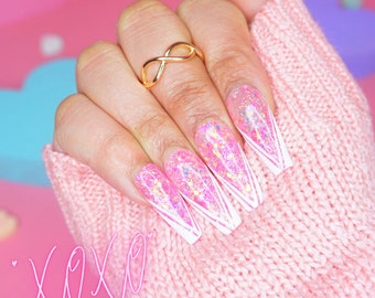 Puede incluir: Uñas largas de stiletto rosa y blanco con purpurina y un diseño de punta blanca. Las uñas se muestran en una mano que lleva un anillo de infinito dorado.