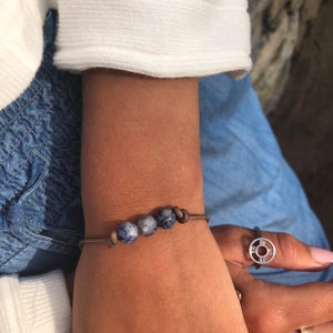 以下が含まれることがあります： 4つの青と白の斑点のあるビーズが付いた茶色の革のブレスレット。ブレスレットは人の手首に巻かれています。その人はローマ数字が刻まれた銀の指輪もしています。