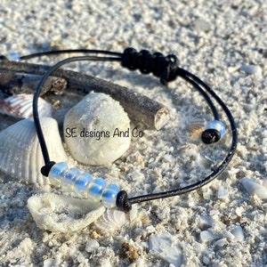 Pode incluir: Um bracelete de cordão de couro preto com uma fila de contas brancas iridescentes. O bracelete é amarrado com um nó em cada extremidade.