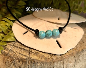 May include: A simple turquoise bead necklace with three beads on a black leather cord. The necklace is displayed on a white seashell.