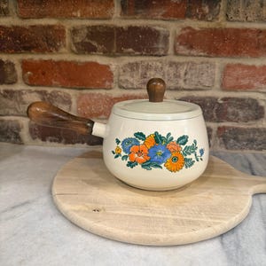 Könnte beinhalten: Cremefarbener Vintage-Kochtopf mit Holzgriff und Deckel. Der Kochtopf hat ein Blumendesign in Orange, Blau und Gelb. Er steht auf einem Holzschneidebrett, was auf die Verwendung in der Küche oder im Esszimmer hindeutet.