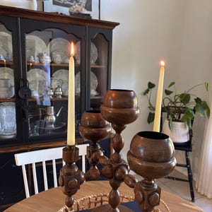 Puede incluir: Un candelabro de madera con tres candeleros y velas encendidas. El candelabro es marrón y tiene un diseño decorativo. Las velas son largas y delgadas. El candelabro está sobre una mesa redonda de madera.