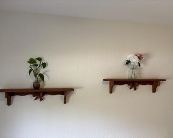 May include: Two wooden shelves with a dark finish are mounted on a light-colored wall. One shelf holds a small plant in a glass vase, and the other holds a bouquet of white and pink flowers in a glass vase. Each shelf has a decorative bow.