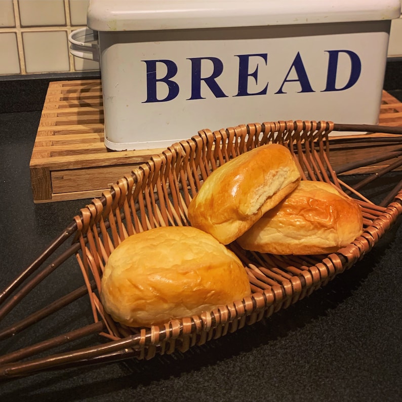 Pode incluir: Tr&ecirc;s p&atilde;es dourados em uma cesta de vime tran&ccedil;ada. A cesta est&aacute; sobre uma superf&iacute;cie preta. Os p&atilde;es est&atilde;o dispostos em semic&iacute;rculo. Uma caixa de p&atilde;o branca com a palavra "BREAD" em letras azuis est&aacute; no fundo.