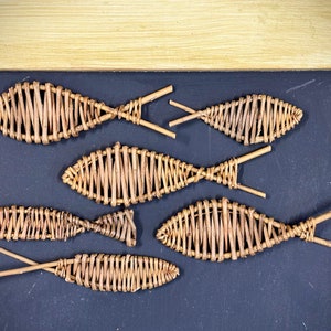 Könnte beinhalten: Ein gerahmtes Kunstwerk mit sieben gewebten Weidenfischen auf dunklem blauem Hintergrund. Der Rahmen ist hellbeige mit Seilakzenten in den Ecken. Die Fische sind unterschiedlich groß und haben stäbchenartige Schwänze.
