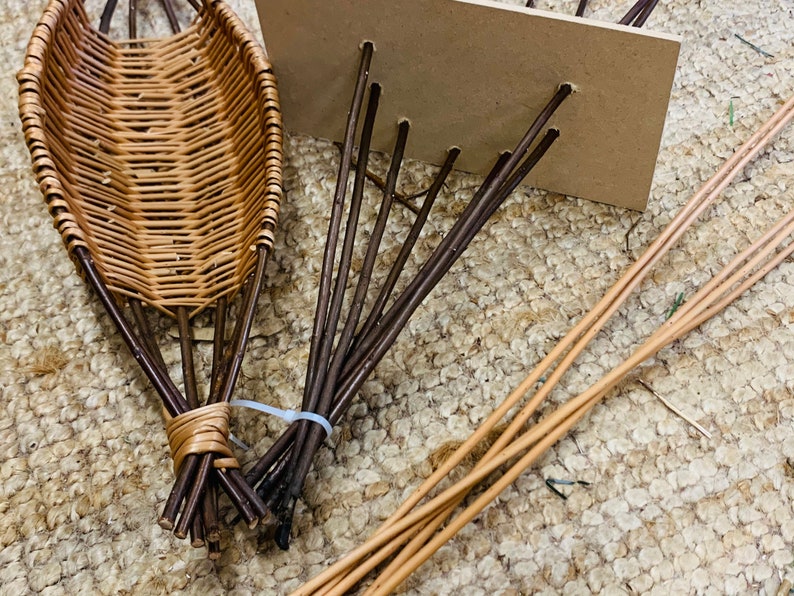 Pode incluir: Uma cesta de vime com fios tecidos castanhos e bege, sendo constru&iacute;da com ramos castanhos amarrados com um lacinho de pl&aacute;stico transparente. A cesta est&aacute; sentada em um tapete bege.