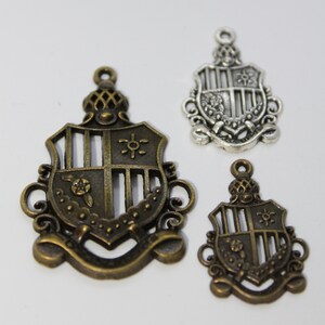 May include: Three decorative shield charms in bronze and silver tones. The charms feature ornate designs with floral and geometric elements. The largest charm is bronze with a detailed crest design. The smallest charm is also bronze, and the middle charm is silver.