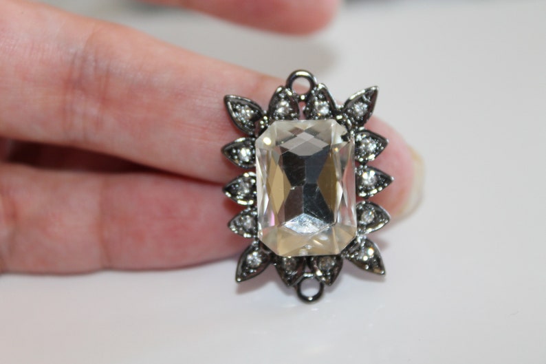 Puede incluir: Un gran cristal transparente facetado est&aacute; engastado en un marco de metal plateado con un dise&ntilde;o floral decorativo. El marco tiene un bucle en la parte superior para colgar.