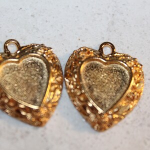 May include: Two gold-tone heart-shaped pendant charms with a filigree design. The charms have a smooth, slightly textured surface and a hollow center.