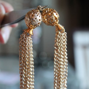 May include: Gold-coloured tassel earrings. Each earring has a filigree ball top, a coiled accent, and a long chain tassel. The earrings are held by a tool, showcasing their intricate design and elegant style.
