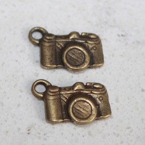 May include: Two antique gold-toned camera charms. The charms are detailed with a lens, shutter button, and other camera features.