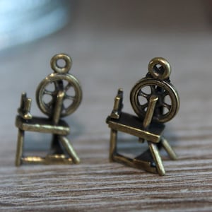 May include: Two antique gold spinning wheel charms with a detailed design. The charms are on a wooden surface.