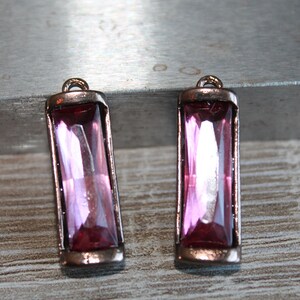 May include: Two rectangular, pink gemstone pendants with a copper-colored metal frame. Each pendant has a loop at the top for hanging. The gemstones are a vibrant pink color and are faceted to catch the light.