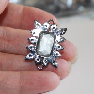 Puede incluir: Pendiente de metal plateado con una piedra rectangular transparente y facetada en el centro. El pendiente est&aacute; rodeado por un dise&ntilde;o decorativo en forma de estrella.