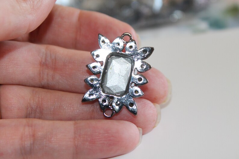 Puede incluir: Pendiente de metal plateado con una piedra rectangular transparente y facetada en el centro. El pendiente est&aacute; rodeado por un dise&ntilde;o decorativo en forma de estrella.