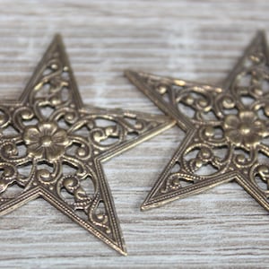 May include: Two ornate gold metal stars with a filigree design. The stars have a flower in the center and are on a wood background.