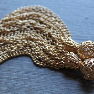 Könnte beinhalten: Nahaufnahme eines goldfarbenen Quastenanhängers. Der Anhänger besteht aus mehreren Strängen kleiner Ketten und zwei dekorativen Filigran-Kugelakzenten. Das Metall hat eine helle, glänzende Oberfläche. Der Hintergrund ist eine dunkelgraue Holzoberfläche.