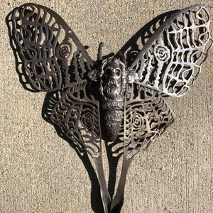 May include: A metal sculpture of a butterfly with intricate cutouts and a human face in the center. The sculpture is painted in a dark metallic color.