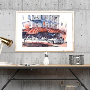 Puede incluir: Una pintura de acuarela de un caf&eacute; parisino con una marquesina a rayas rojas y blancas, gente sentada afuera y un coche antiguo estacionado en frente. La pintura est&aacute; enmarcada y colgada en una pared de concreto sobre un escritorio de madera con una l&aacute;mpara, un libro y un jarr&oacute;n de l&aacute;pices.