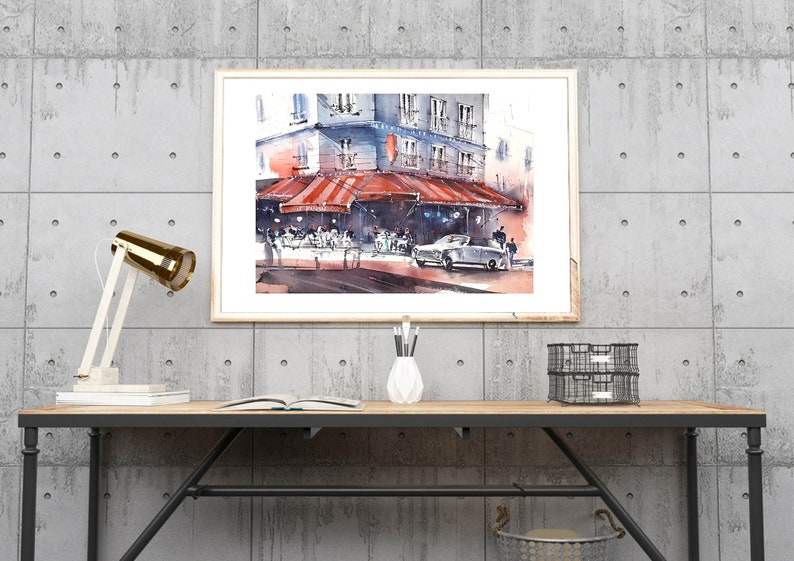 Puede incluir: Una pintura de acuarela de un caf&eacute; parisino con una marquesina a rayas rojas y blancas, gente sentada afuera y un coche antiguo estacionado en frente. La pintura est&aacute; enmarcada y colgada en una pared de concreto sobre un escritorio de madera con una l&aacute;mpara, un libro y un jarr&oacute;n de l&aacute;pices.
