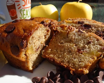 Könnte beinhalten: Nahaufnahme eines Bananenbrotes mit Schokoladenstückchen, geschnitten und bereit zum Essen. Das Brot liegt auf einem weißen Teller mit verstreuten Schokoladenstückchen. Im Hintergrund befinden sich zwei gelbe Äpfel.