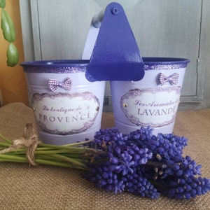 Peut inclure: Deux pots de fleurs en métal de couleur lavande avec une poignée bleue. Les pots ont un design décoratif avec des nœuds et du texte en français. Le texte sur le pot de gauche est "La boutique de Provence" et le texte sur le pot de droite est "Les Aromates de LAVANDE". Un bouquet de fleurs violettes se trouve devant les pots.