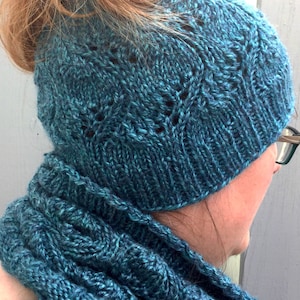 May include: A teal blue knitted beanie hat featuring a delicate lace-like leaf pattern and a ribbed brim, paired with a matching teal blue knitted scarf that displays a textured, repeating cable-knit design throughout the fabric.