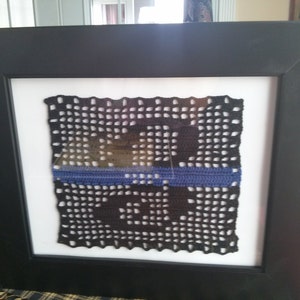 May include: A framed black and white crocheted square with a blue stripe in the middle. The crocheted square is in a black frame.