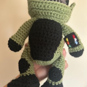May include: Green and black crocheted plush toy with a gray head and a rainbow detail on one arm. The toy is in the shape of a person and has a rounded body.