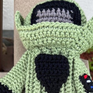 May include: A handmade crochet Yoda doll in shades of green, black, and gray. The doll features large ears, a black chest, and a control panel detail. The crochet work is visible, showcasing the texture of the yarn.