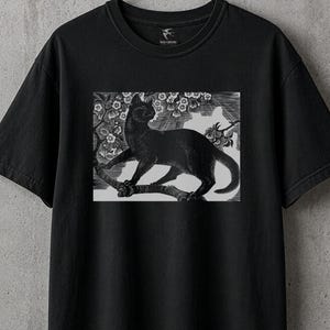 May include: Black t-shirt featuring a black and white illustration of a black cat perched on a tree branch with flowers. The cat is the focal point of the design, set against a backdrop of floral elements.