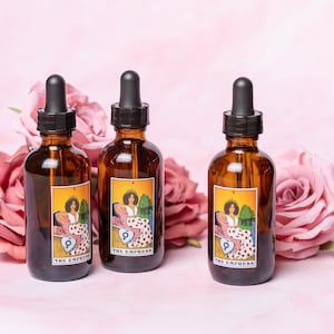 May include: Three brown glass bottles with droppers, each with a label featuring a tarot card image of "The Empress". The bottles are arranged in a row on a pink background with pink roses.