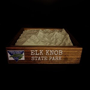 May include: A wooden framed 3D topographic map of Elk Knob State Park in North Carolina. The map is made of a light brown material and is set in a dark brown wooden frame. The frame has a white label that reads "Elk Knob State Park" and a smaller label that reads "North Carolina State Parks" with a blue and white logo.