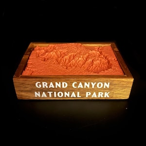 May include: A 3D printed model of the Grand Canyon National Park in a wooden frame. The model is a light orange color and the frame is a dark brown color. The text "Grand Canyon National Park" is printed in white on the front of the frame.