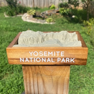 May include: A wooden box with a 3D model of Yosemite National Park inside. The box is brown and has the words "Yosemite National Park" printed on the front in white.
