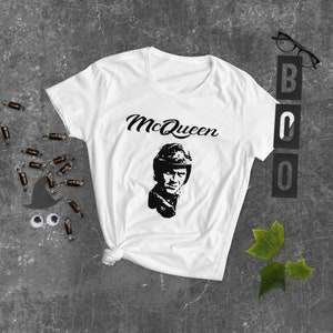 May include: White t-shirt with a black and white image of Steve McQueen wearing a helmet and the text "McQueen" in a black cursive font.